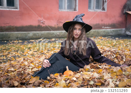 Teen girl in a witch costume sits on a pile of yellow leaves with a small pumpkin nearby, against the backdrop of an old pink wall Teen girl in a witch costume sits on a pile of yellow leaves with a small pumpkin nearby, against the backdrop of an old pink wall 132511293