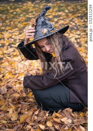 Smiling teen girl in a witch hat and brown jacket plays among fallen autumn leaves, creating a joyful Halloween atmosphere Smiling teen girl in a witch hat and brown jacket plays among fallen autumn leaves, creating a joyful Halloween atmosphere 132511294