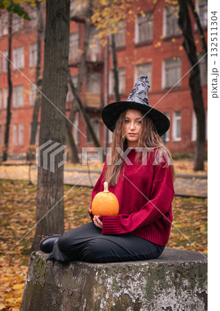 Teen girl in burgundy sweater and witch hat holds a pumpkin outdoors, surrounded by autumn leaves and brick buildings, creating a cozy Halloween vibe 132511304