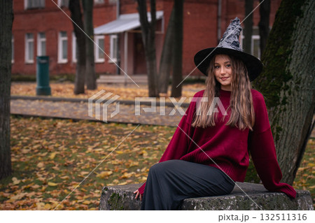 Teen girl in a witch hat and burgundy sweater sits on a stone surface surrounded by autumn leaves and trees near brick buildings 132511316