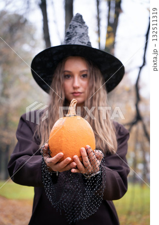 Teen girl in a witch hat holds an orange pumpkin forward, with a soft focus background of autumn trees creating a mystical Halloween mood Teen girl in a witch hat holds an orange pumpkin forward, with a soft focus background of autumn trees creating a mystical Halloween mood 132511319