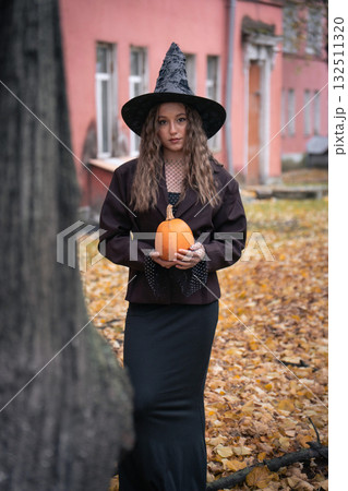 Teen girl in a witch hat and dark outfit holds an orange pumpkin outdoors, with autumn trees and pink brick buildings in the background Teen girl in a witch hat and dark outfit holds an orange pumpkin outdoors, with autumn trees and pink brick buildings in the background 132511320