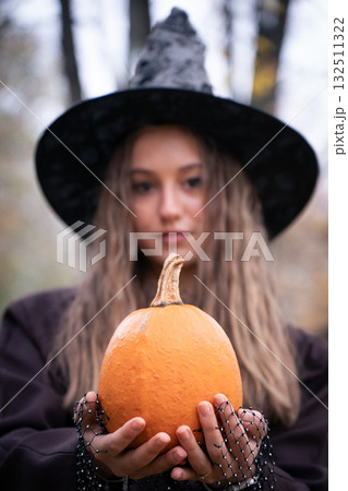 Teen girl in a witch hat holds an orange pumpkin forward, with a soft focus background of autumn trees creating a mystical Halloween mood Teen girl in a witch hat holds an orange pumpkin forward, with a soft focus background of autumn trees creating a mystical Halloween mood 132511322