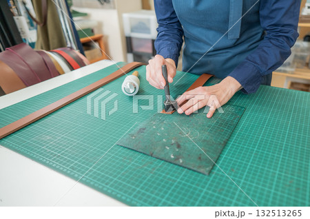 Leatherworker cutting a piece of leather from a belt in a workshop.  132513265