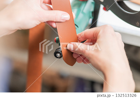Leatherworker grinding the edge of a strip of leather for a belt in the workshop.  132513326