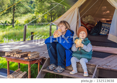 Toddler and his teenage brother tourists in warm vests and hats eating buns while sitting on glamping steps, enjoying family travel, outdoor adventure, and bonding 132513638