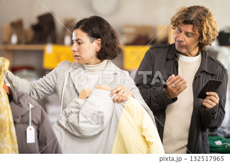 Woman choose jacket in store, bored husband accompanies his wife during shopping Woman choose jacket in store, bored husband accompanies his wife during shopping 132517965