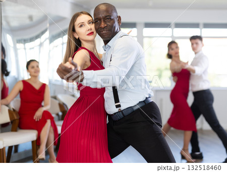 Smiling young european woman in red clothes dancing latin dance with African american man in elegant clothes, energetic and fast tango in dance salon 132518460