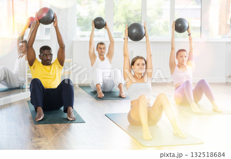 Pilates class in multi-ethnic group - men and women hold the ball above their heads and do bends Pilates class in multi-ethnic group - men and women hold the ball above their heads and do bends 132518684
