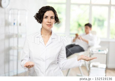 Middle-aged woman doctor posing in doctor's cabinet with patient lying on clinical chair 132519136