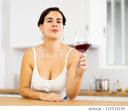 Thoughtful young hispanic woman relaxing in kitchen with glass of wine 132519139