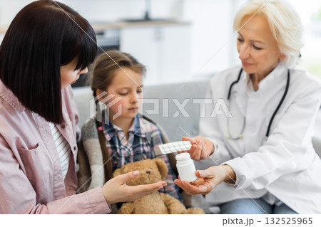 A doctor is showing medication to a young girl and her mother. The child is wrapped in a blanket, appearing unwell, while the doctor explains the treatment. A doctor is showing medication to a young girl and her mother. The child is wrapped in a blanket, appearing unwell, while the doctor explains the treatment. 132525965
