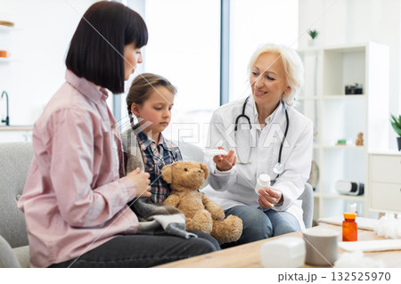 A doctor is showing medication to a mother and her daughter, who is wrapped in a blanket and holding a teddy bear, likely at a clinic. 132525970