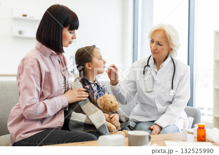 A doctor checks a young girl's temperature while the mother comforts her. The girl is wrapped in a blanket and holding a teddy bear. A doctor checks a young girl's temperature while the mother comforts her. The girl is wrapped in a blanket and holding a teddy bear. 132525990