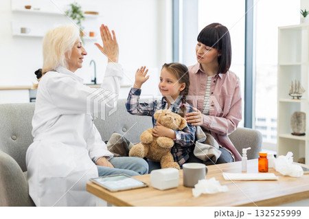A doctor gives a high five to a young girl, who is sitting with her mother on a sofa, possibly after a checkup. The girl holds a teddy bear. A doctor gives a high five to a young girl, who is sitting with her mother on a sofa, possibly after a checkup. The girl holds a teddy bear. 132525999