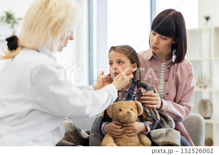 A doctor checks a young girl's throat while her mother comforts her. The girl is wrapped in a blanket, holding a teddy bear. 132526002