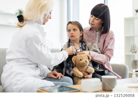 A female doctor uses a stethoscope to listen to a young girl's heart while her mother comforts her. The girl holds a teddy bear. A female doctor uses a stethoscope to listen to a young girl's heart while her mother comforts her. The girl holds a teddy bear. 132526008