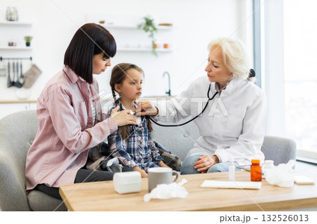 A doctor uses a stethoscope to examine a young girl while her mother sits beside her, providing comfort and support during the checkup. A doctor uses a stethoscope to examine a young girl while her mother sits beside her, providing comfort and support during the checkup. 132526013