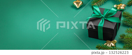 Black gift box with green satin ribbon and fir branches on green background, elegant Christmas composition with golden ornaments and copy space for text, festive flat lay 132526223