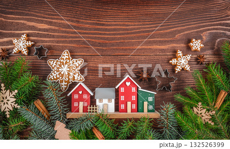 Cozy Christmas village scene with decorative houses, gingerbread snowflakes, fir branches, and festive spices on a rustic wooden background. copy space. top view 132526399