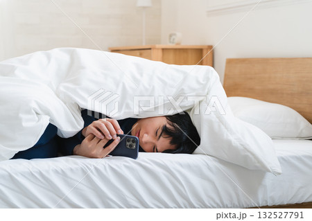 布団にくるまりスマートフォンを操作している若い男性　ライフスタイルイメージ 132527791