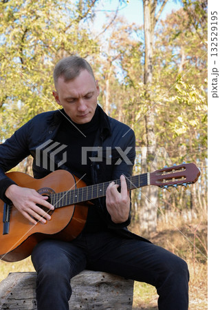 Portrait of smiling caucasian artist plays wooden acoustic guitar in autumnal park. Young songwriter plays string musical instrument hobby outside in nature fall time. Audio music healing Portrait of smiling caucasian artist plays wooden acoustic guitar in autumnal park. Young songwriter plays string musical instrument hobby outside in nature fall time. Audio music healing 132529195