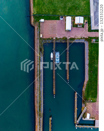 Aerial view of marina two boats docked between parallel wooden piers in green blue water, bordered by rocky shore, red roofed building 132531220