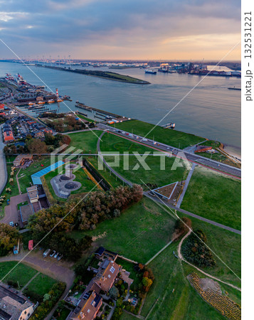 Aerial view of river port at sunset cargo ships, cranes, and industrial docks line both banks, with dense urban grid and green fields framing the waterway in a dynamic commercial landscape. 132531221