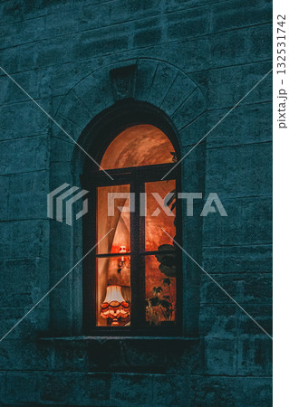 The image shows an arched window set into a stone wall during twilight or night time. The window is lit from the inside, casting a warm glow. Through the pane, one can see indoor plants and what looks The image shows an arched window set into a stone wall during twilight or night time. The window is lit from the inside, casting a warm glow. Through the pane, one can see indoor plants and what looks 132531742