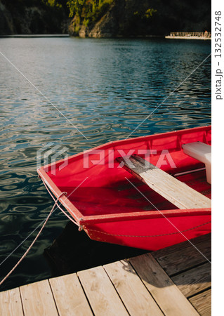 Red rowboat with wooden seats gently rocking on calm lake waters, secured to wooden dock, creating serene and inviting atmosphere perfect for relaxation and leisure. Wooden red rowboat floating 132532748