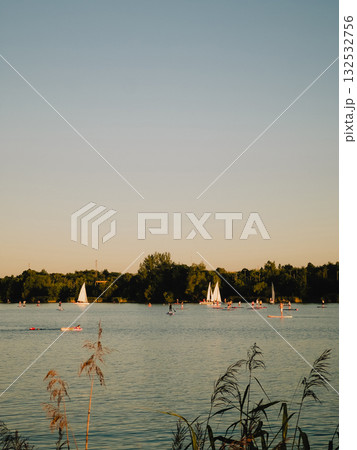 Sailboats gliding across calm lake and paddle boarders enjoying golden hour create picturesque scene, framed by lush vegetation and clear, vibrant sky. Sailboats gliding across calm lake waters Sailboats gliding across calm lake and paddle boarders enjoying golden hour create picturesque scene, framed by lush vegetation and clear, vibrant sky. Sailboats gliding across calm lake waters 132532756