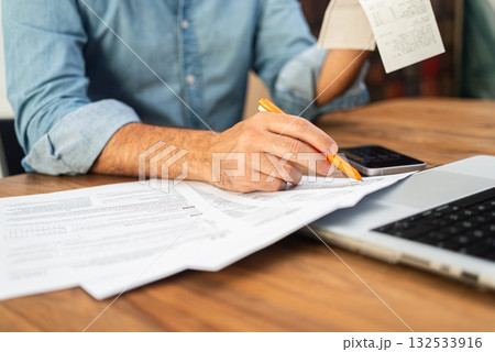 Man in a denim shirt calculating taxes with receipts and forms on a wooden desk. Tax preparation and financial management concept.  132533916