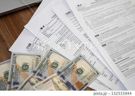 Tax forms and dollar bills spread out on a wooden desk near a laptop. Financial management, tax refund, and accounting concept. Tax forms and dollar bills spread out on a wooden desk near a laptop. Financial management, tax refund, and accounting concept. 132533944