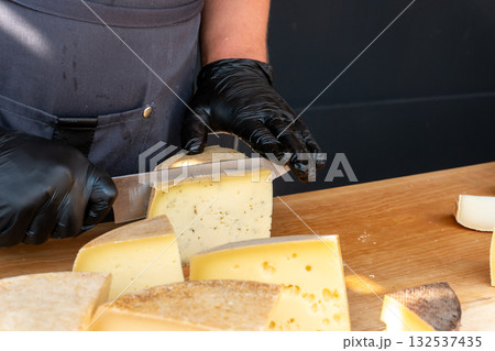 A vendor slices a piece of cheese with a knife at a farmers market A vendor slices a piece of cheese with a knife at a farmers market 132537435