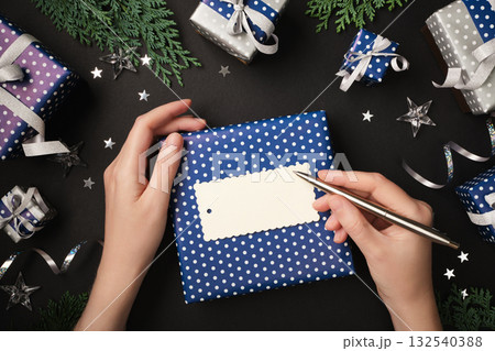 Hands sign a gift tag. Ready-made gifts, ribbon, and thuja twigs surround it. Copy space. Hands sign a gift tag. Ready-made gifts, ribbon, and thuja twigs surround it. Copy space. 132540388