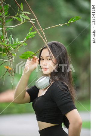 Young woman with headphones posing gracefully outdoors in a lush green setting, showcasing her natural beauty and confidence in a casual fashion style 132541164