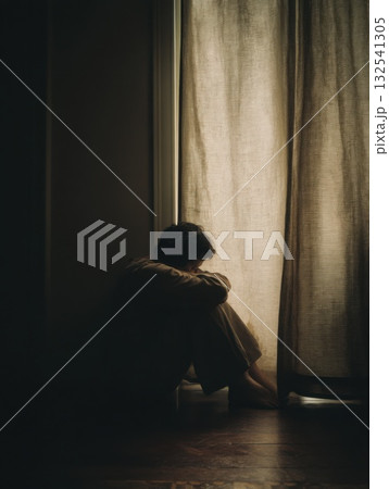 Silhouetted figure sits alone by a window in low light. Soft curtain, shadows, and quiet emotion convey solitude, reflection, and mental stillness 132541305