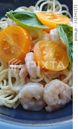Close-up of creamy pasta with shrimp, yellow cherry tomatoes, and fresh basil leaves served on a blue plate. The image represents Italian cuisine, gourmet seafood, and Mediterranean flavors 132541571