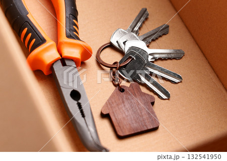 Curved nose pliers and bunch of door keys with wooden house keychain inside cardboard box. Symbol of home repair and DIY 132541950