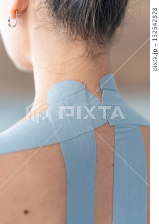Close-up view of kinesiology tape applied to the neck and shoulder area of a woman. Physical therapy and muscle support through modern rehabilitation techniques Close-up view of kinesiology tape applied to the neck and shoulder area of a woman. Physical therapy and muscle support through modern rehabilitation techniques 132542878