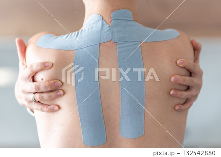 Young woman with kinesiology tape on her back and shoulders, embracing herself with both hands. Physical therapy, body awareness and emotional self-care through healing touch 132542880