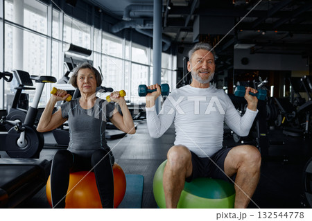 Mature couple sit on exercise balls in a gym with dumbbells Mature couple sit on exercise balls in a gym with dumbbells 132544778