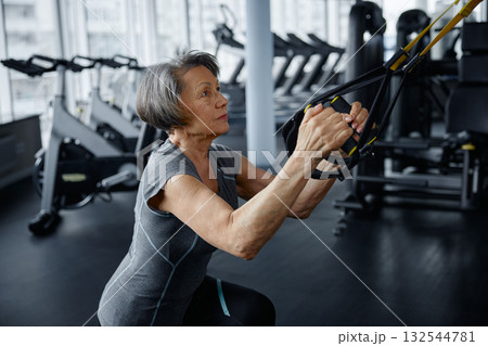 An elderly woman is exercising on a suspension trainer in a gym 132544781