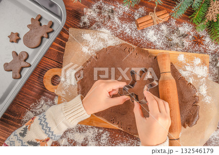 Hands cutting gingerbread dough with cookie cutter on wooden board, Christmas baking process with flour, rolling pin, cinnamon sticks, fir branches, and baking tray, cozy holiday atmosphere, top view. 132546179