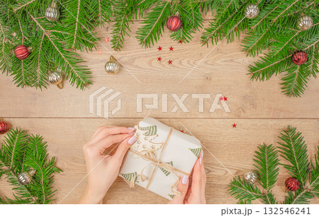 Woman wrapping Christmas gift with fir branches and festive ornaments on wooden background, top view with copy space, holiday preparation and handmade present concept 132546241