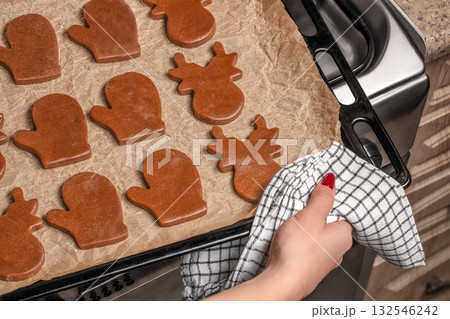 Homemade gingerbread cookies on baking tray with parchment paper, womans hand placing tray into oven, holiday baking preparation in cozy kitchen, top view close-up 132546242