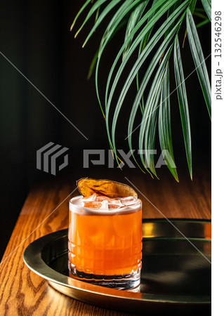 Vibrant orange cocktail with ice cubes and dried pear garnish served on a brass tray, illuminated by warm light and tropical leaves in the background Vibrant orange cocktail with ice cubes and dried pear garnish served on a brass tray, illuminated by warm light and tropical leaves in the background 132546298
