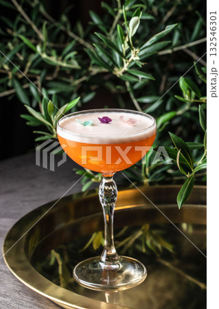 Elegant orange cocktail with foam and edible flower petals, served in a crystal coupe glass on a golden tray, surrounded by green leaves 132546301