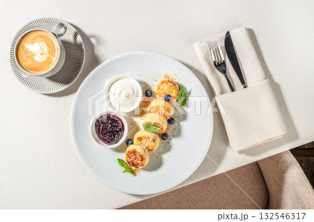 Breakfast table with cottage cheese pancakes, sour cream, blueberry jam, and a cup of cappuccino served in a cozy cafe setting, top view 132546317