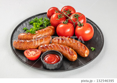 Juicy grilled sausages served with fresh tomatoes, parsley, and ketchup on a black plate over light background 132546395
